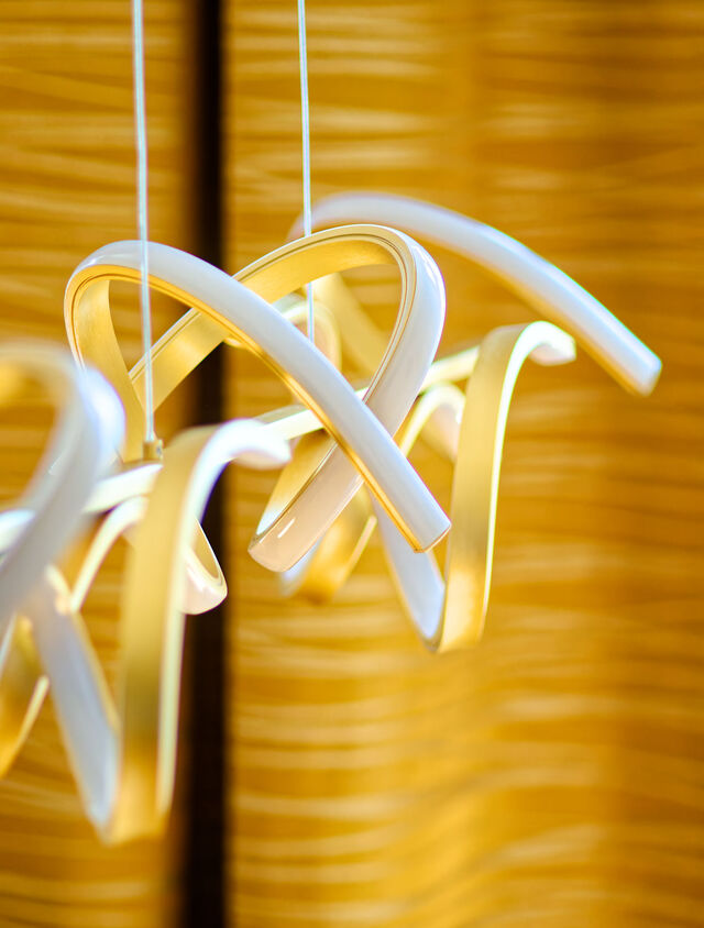Nahaufnahme moderner skulpturaler Pendelleuchte mit verschlungenen gold-wei&szlig;en Metallringen &uuml;ber Essbereich im Premium-m&ouml;blierten Apartment Bamberg