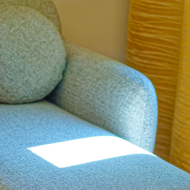 Nahaufnahme Designer-Sofa mit hochwertigem grau-meliertem Salz-und-Pfeffer-Polsterstoff, warmem Sonnenfleck und goldenen Vorh&auml;ngen im Hintergrund im m&ouml;blierten Apartment Bamberg