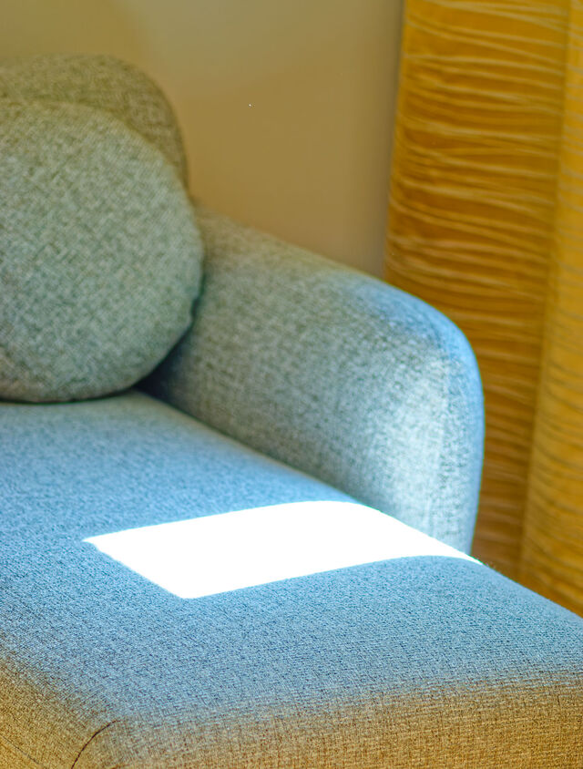 Nahaufnahme Designer-Sofa mit hochwertigem grau-meliertem Salz-und-Pfeffer-Polsterstoff, warmem Sonnenfleck und goldenen Vorh&auml;ngen im Hintergrund im m&ouml;blierten Apartment Bamberg