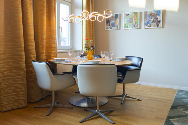 Moderner Essbereich mit rundem Tisch, sechs wei&szlig;en St&uuml;hlen, goldenen Vorh&auml;ngen und drei Wandbildern in hellem Raum mit Holzboden.
