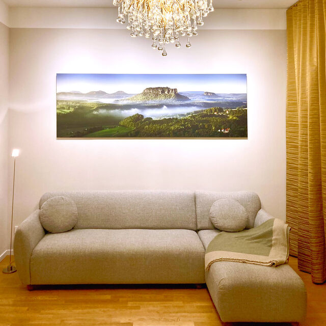 Abendansicht Wohnzimmer mit grauem Salz-und-Pfeffer-Sofa, gro&szlig;formatigem Landschaftsfoto Lilienstein III von Dirk Wiedlein, Kristall-Icicle-Kronleuchter von Paul Neuhaus und goldenen Zimmer & Rohde Vorh&auml;ngen im m&ouml;blierten Apartment Bamberg