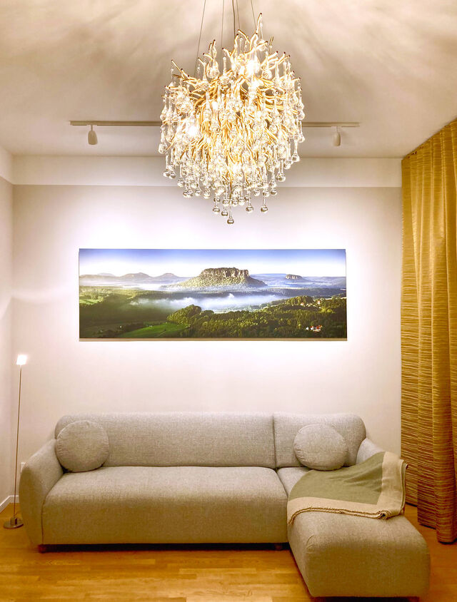 Abendansicht Wohnzimmer mit grauem Salz-und-Pfeffer-Sofa, gro&szlig;formatigem Landschaftsfoto Lilienstein III von Dirk Wiedlein, Kristall-Icicle-Kronleuchter von Paul Neuhaus und goldenen Zimmer & Rohde Vorh&auml;ngen im m&ouml;blierten Apartment Bamberg