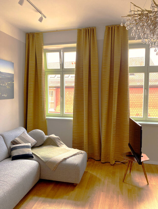 Sonniges Wohnzimmer mit grauem Salz-und-Pfeffer-Sofa, Dekokissen, 45-Zoll Metz HD-Fernseher auf Holz-Retro-St&auml;nder, Paul Neuhaus Icicle-Kronleuchter, goldenen Zimmer & Rohde Vorh&auml;ngen und Blick auf rote Ziegeld&auml;cher im m&ouml;blierten Apartment Bamberg